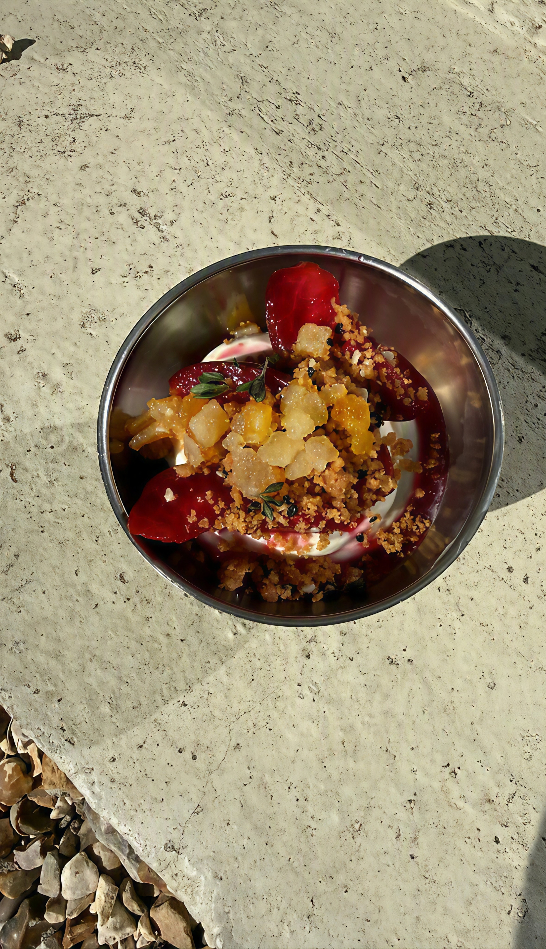 a metal bowl of dessert with poached fruit, crumb and thyme on a sunlit stone surface